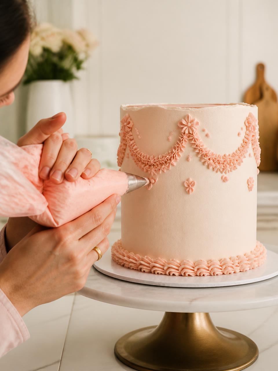 Cake artist piping delicate blush buttercream detail onto a tall cake